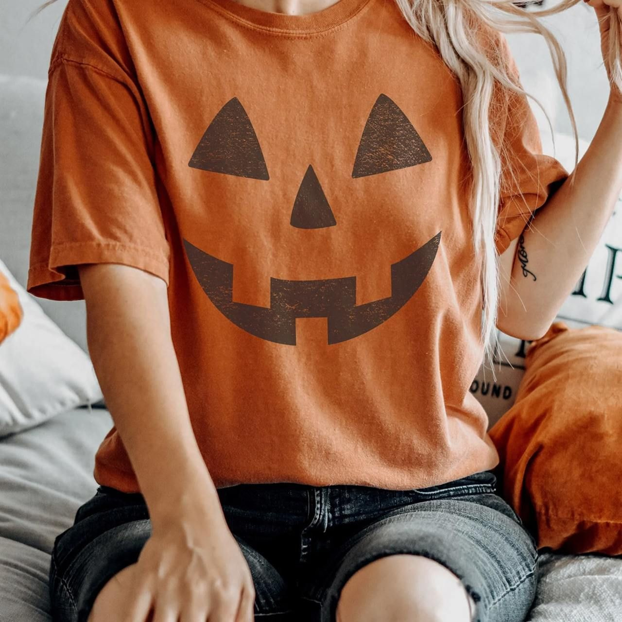 A woman in a Halloween-themed pumpkin face orange t-shirt and denim shorts sits on a couch.